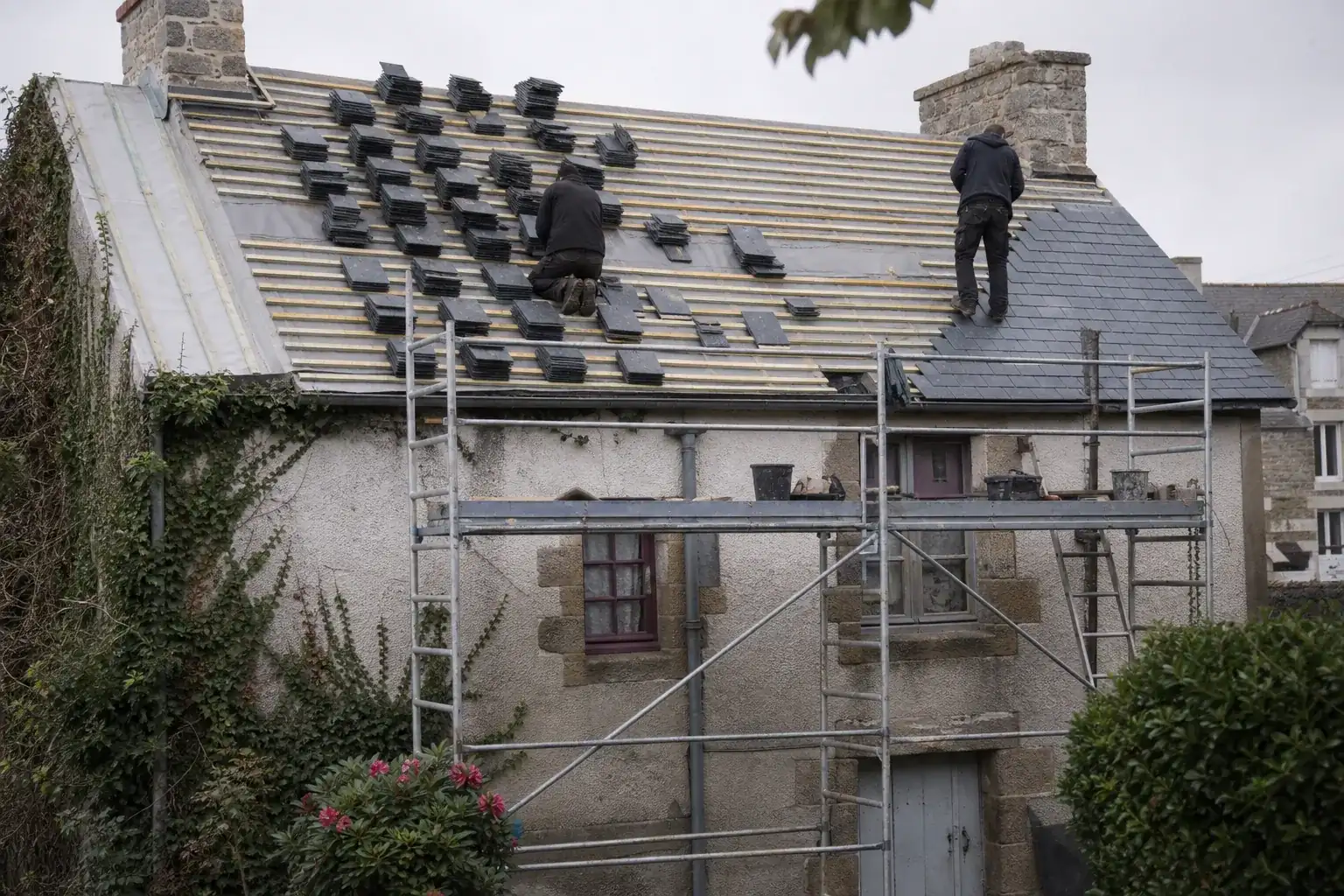 Échafaudage dressé contre une façade enduite, bottes d’ardoise empilées sur les liteaux et écran de sous-toiture à demi déroulé