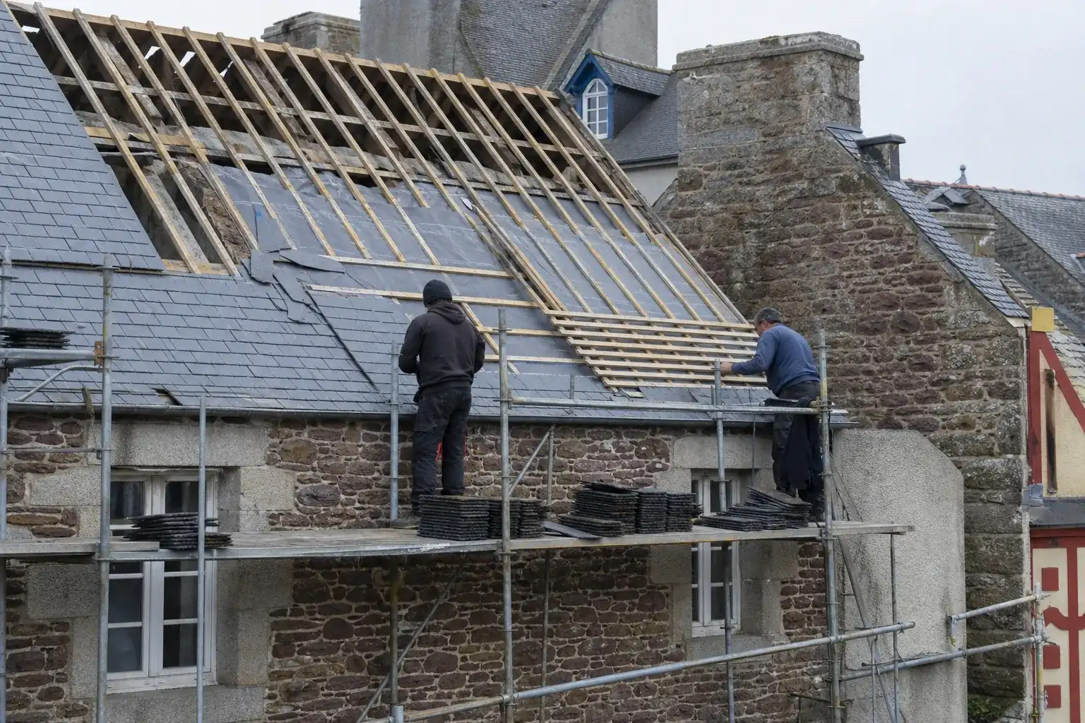 Toiture déposée jusqu’aux chevrons sur une maison de pierre à étage, ardoises récupérées empilées sur la plateforme d’échafaudage